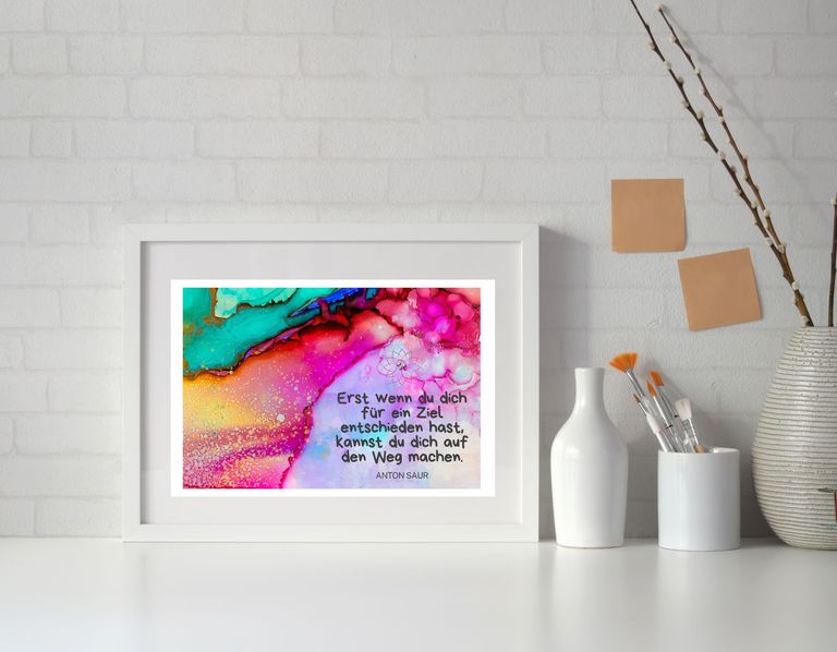 Ein weißer Bilderrahmen auf einem weißen Regal vor einer weißen Backsteinwand mit einem bunten abstrakten Bild und einem deutschen Zitat von Anton Saur. Daneben stehen weiße Behälter mit Pinsel und Stäben sowie eine Vase mit Zweigen.